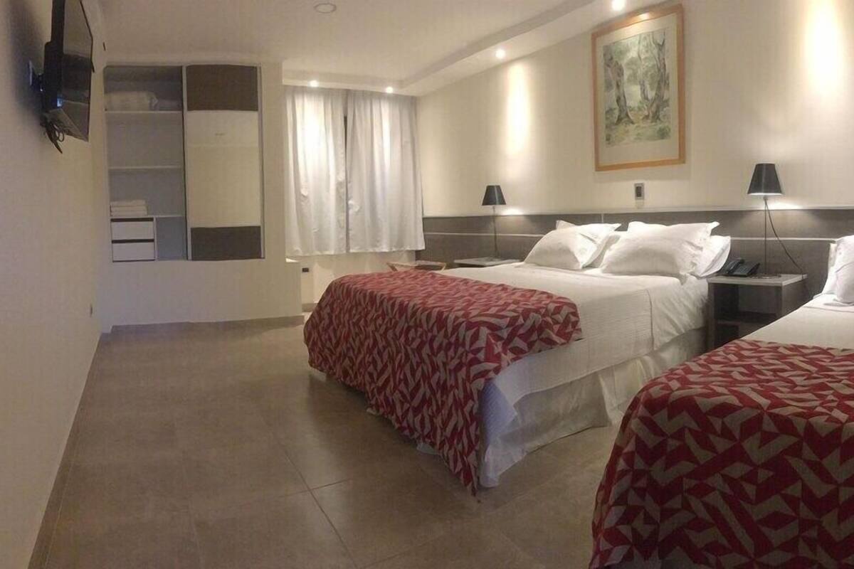 Een hotelkamer met twee bedden, een groot raam met gordijnen, een schilderij aan de muur en een aan de muur bevestigde televisie.