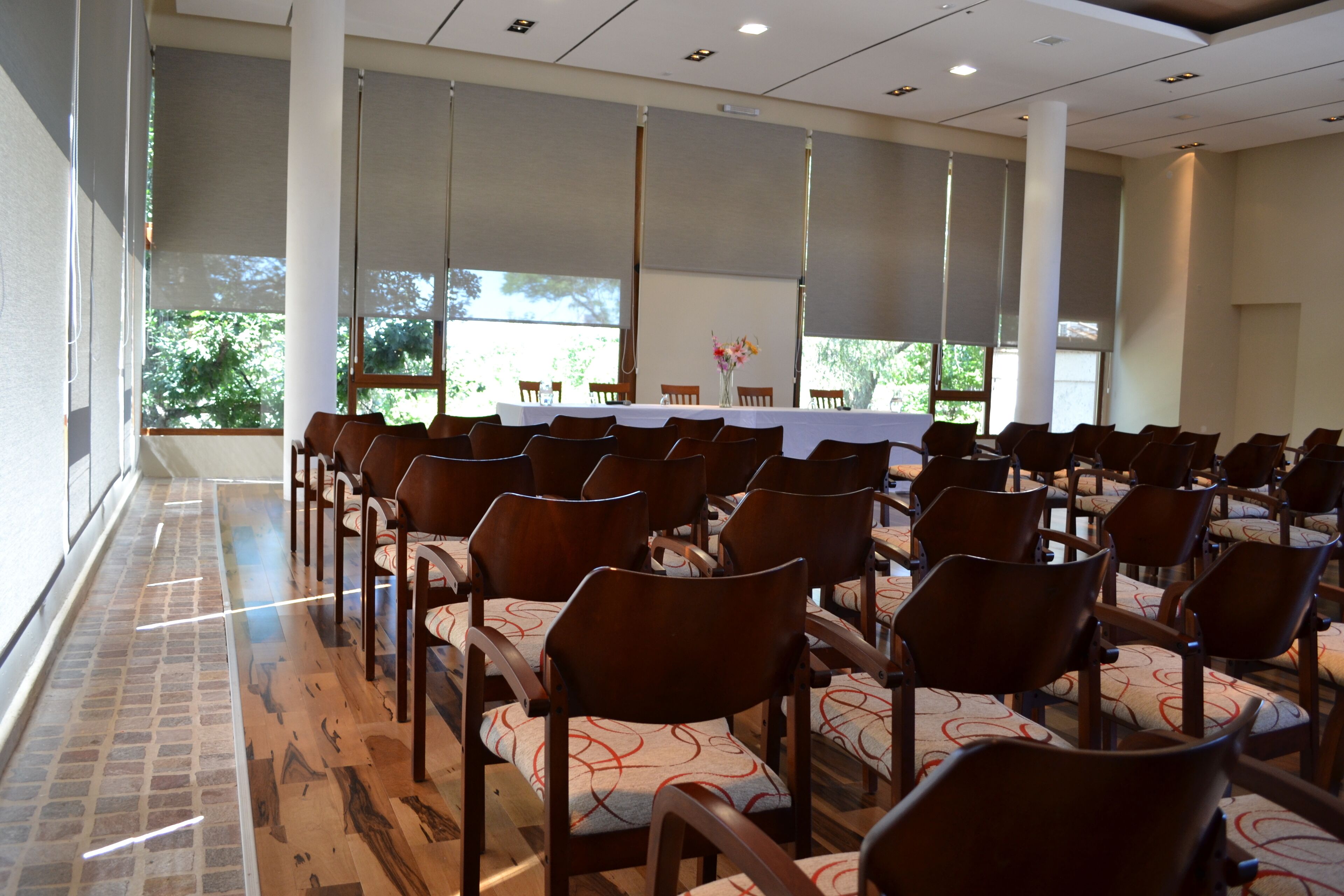 A conference room with rows of wooden chairs and a table, large windows, and a brick floor.