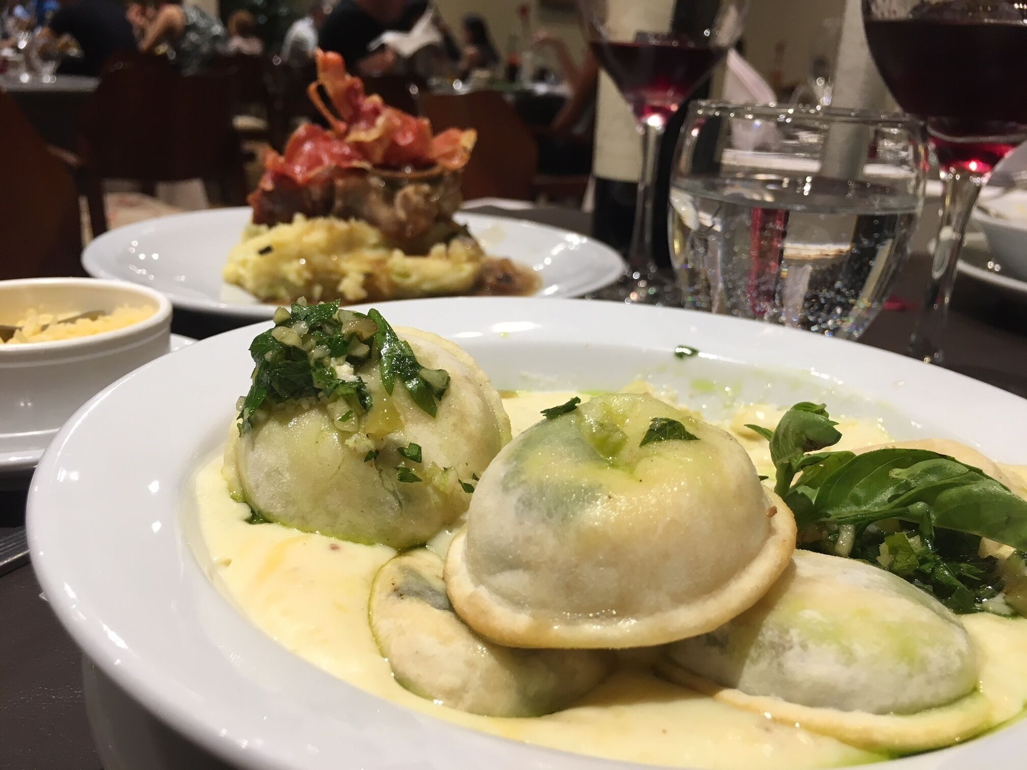 A dining table with a plate of dumplings in a creamy sauce, a glass of red wine, and a bowl of bacon.
