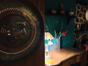 A wooden wall with a decorative plate featuring a figure, a small table with a lamp, and a stone wall with a shelf holding pottery.