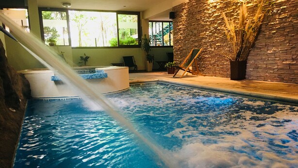 An indoor swimming pool with a water slide, stone wall, and seating area. - Los Espinillos Hotel & Spa (Villa Carlos Paz)