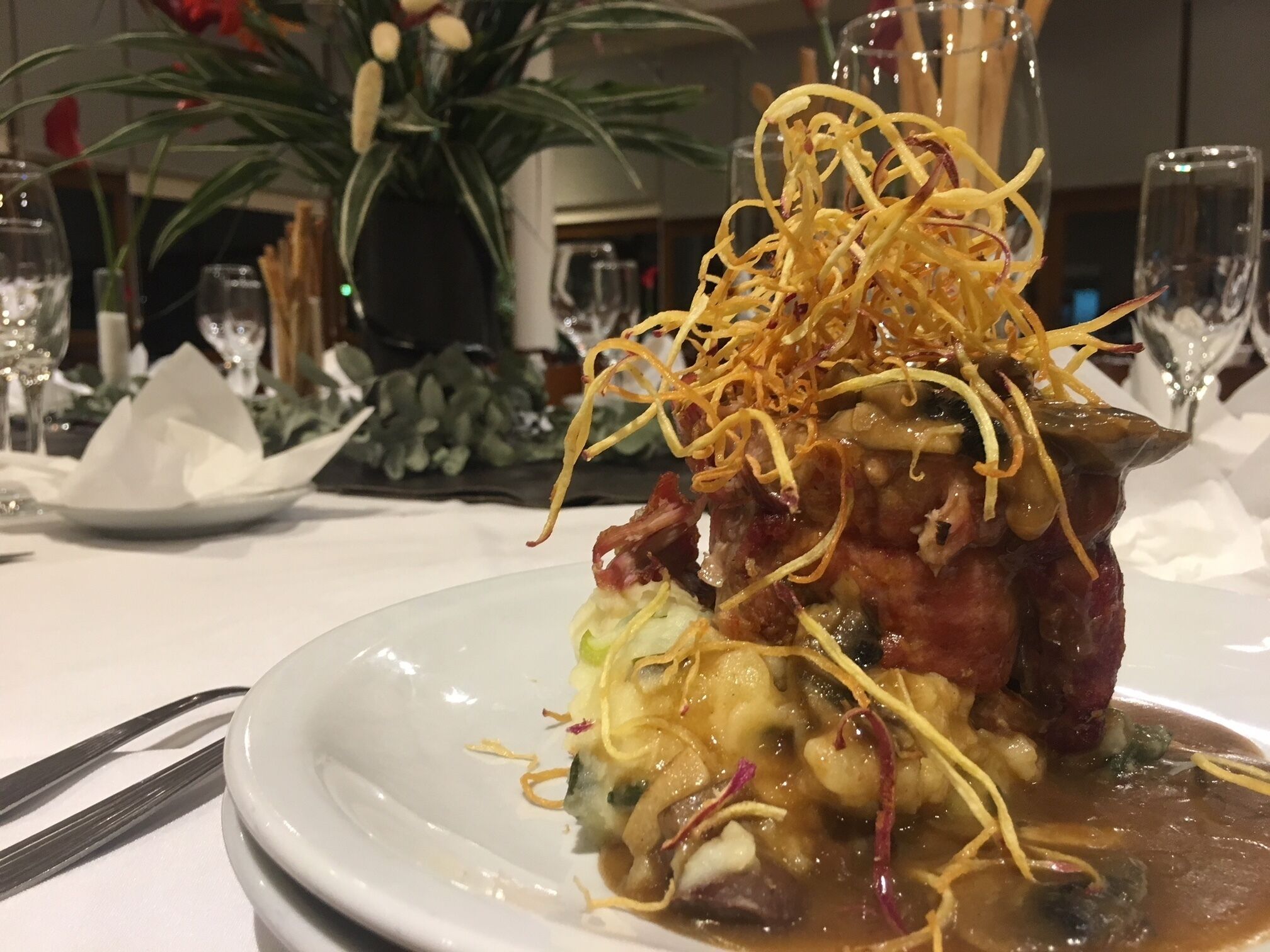 Un plato con guarnición decorativa, un vaso de agua y un jarrón con flores al fondo.