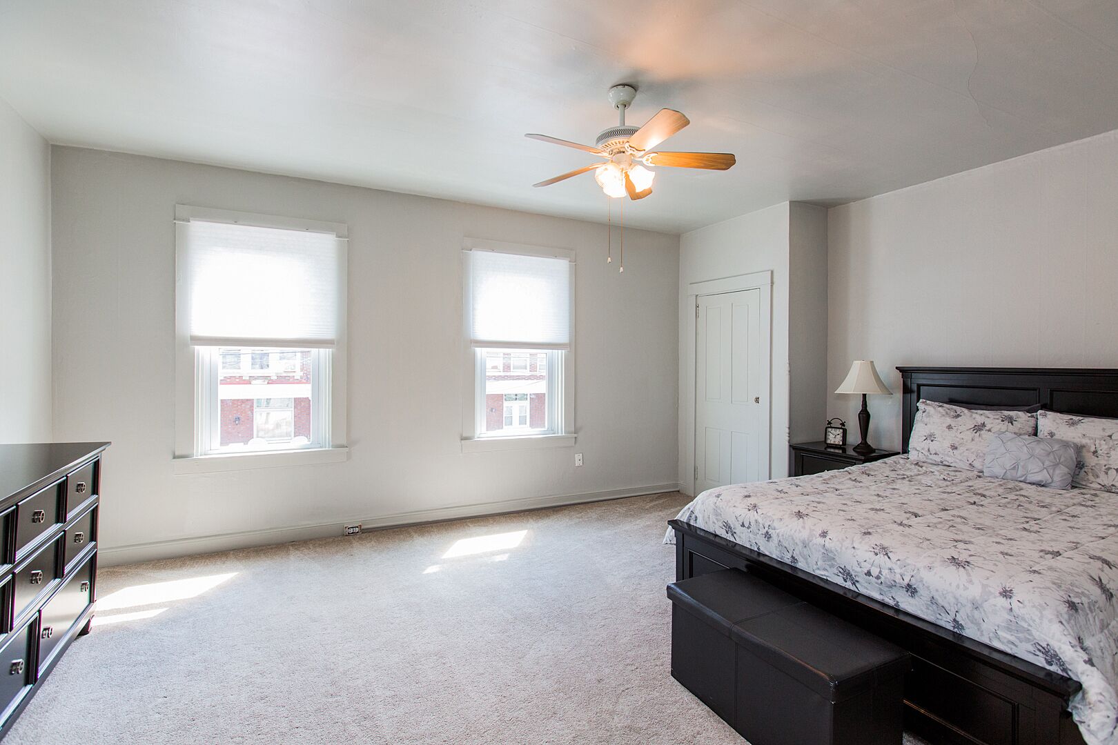 A bedroom with a bed, a nightstand, a dresser, two windows, a ceiling fan, and a door.