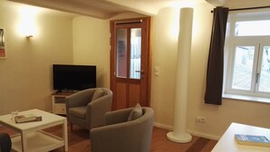 Un salon avec un téléviseur, deux fauteuils, une table basse et une fenêtre avec des rideaux.