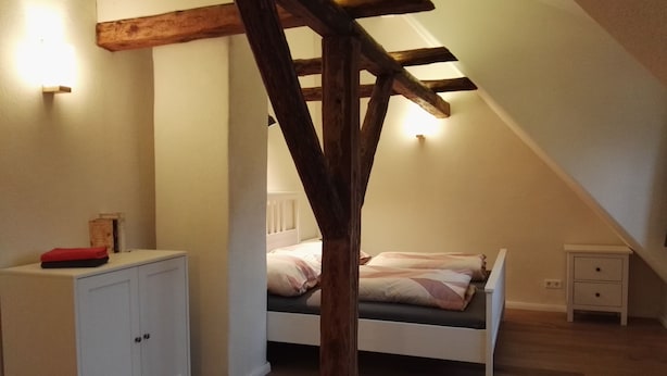 Une chambre avec un plafond en pente, une structure en poutres apparentes, un lit avec des oreillers, une table de chevet et une armoire.