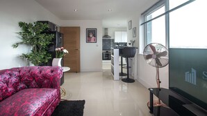 A modern living room with a sofa, a TV, a fan, and a kitchen in the background. - Smart Apartments - The Queens (Southampton)