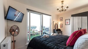 A hotel room with a bed, a TV, a fan, a mirror with lights, and a view of the city. - Smart Apartments - The Queens (Southampton)