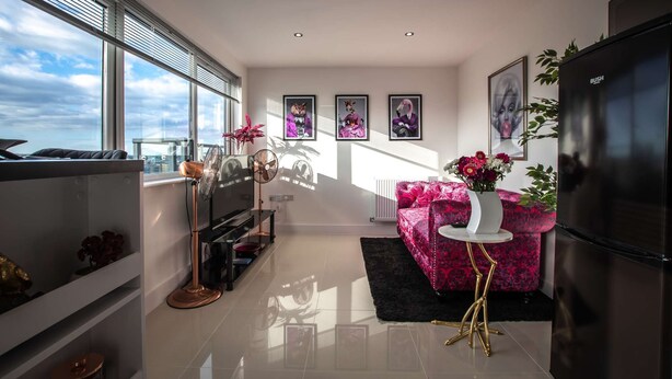 A modern living room with a large window, a pink sofa, a black refrigerator, and a black coffee table. - Smart Apartments - The Queens (Southampton)
