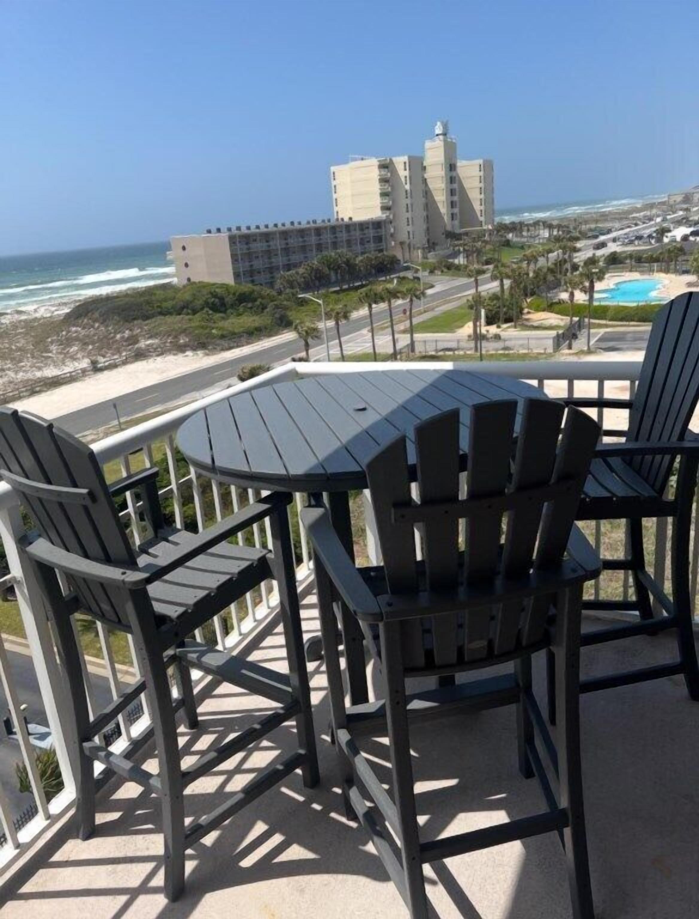 A balcony with Adirondack chairs and a table overlooking a beach and ocean.