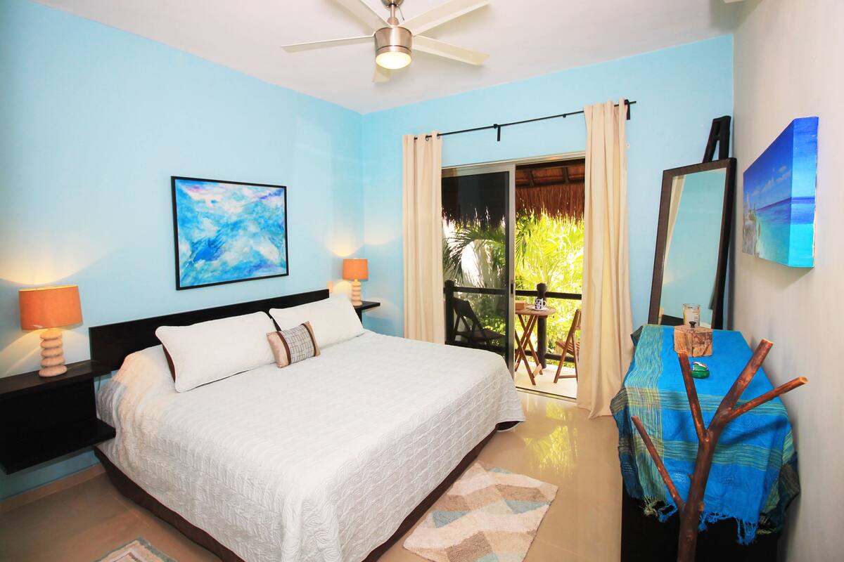 Une chambre avec un lit, un ventilateur de plafond, une table de chevet avec une lampe, un tableau, un balcon avec vue et une table avec une chaise.