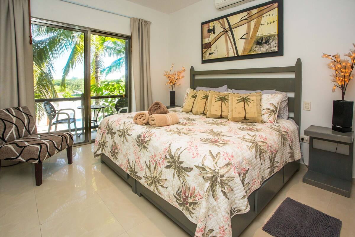 Une chambre d’hôtel avec un lit, une chaise, une petite table, un téléviseur et un balcon donnant sur des palmiers.