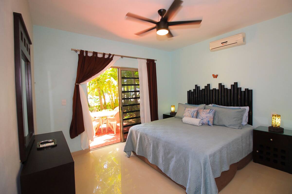 Une chambre à coucher comprenant un lit, un ventilateur de plafond, un appareil de climatisation, une table de chevet et une fenêtre avec des rideaux.