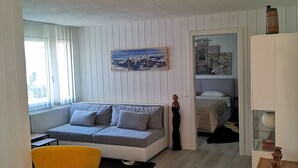 Una sala moderna con un sofá, un sillón amarillo, una mesa de vidrio y una cama al fondo.