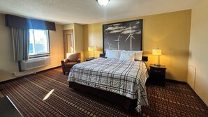 Habitación de hotel con una cama grande, una silla, dos mesitas de noche con lámparas, una ventana con cortinas, un aire acondicionado de pared y un cuadro de aerogeneradores.
