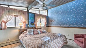 A bedroom with a bed, a ceiling fan, a window with curtains, a wall with floral wallpaper, and a small table with a lamp.