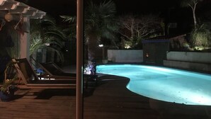 A nighttime view of a swimming pool area with a curved design, surrounded by palm trees and a wooden deck.