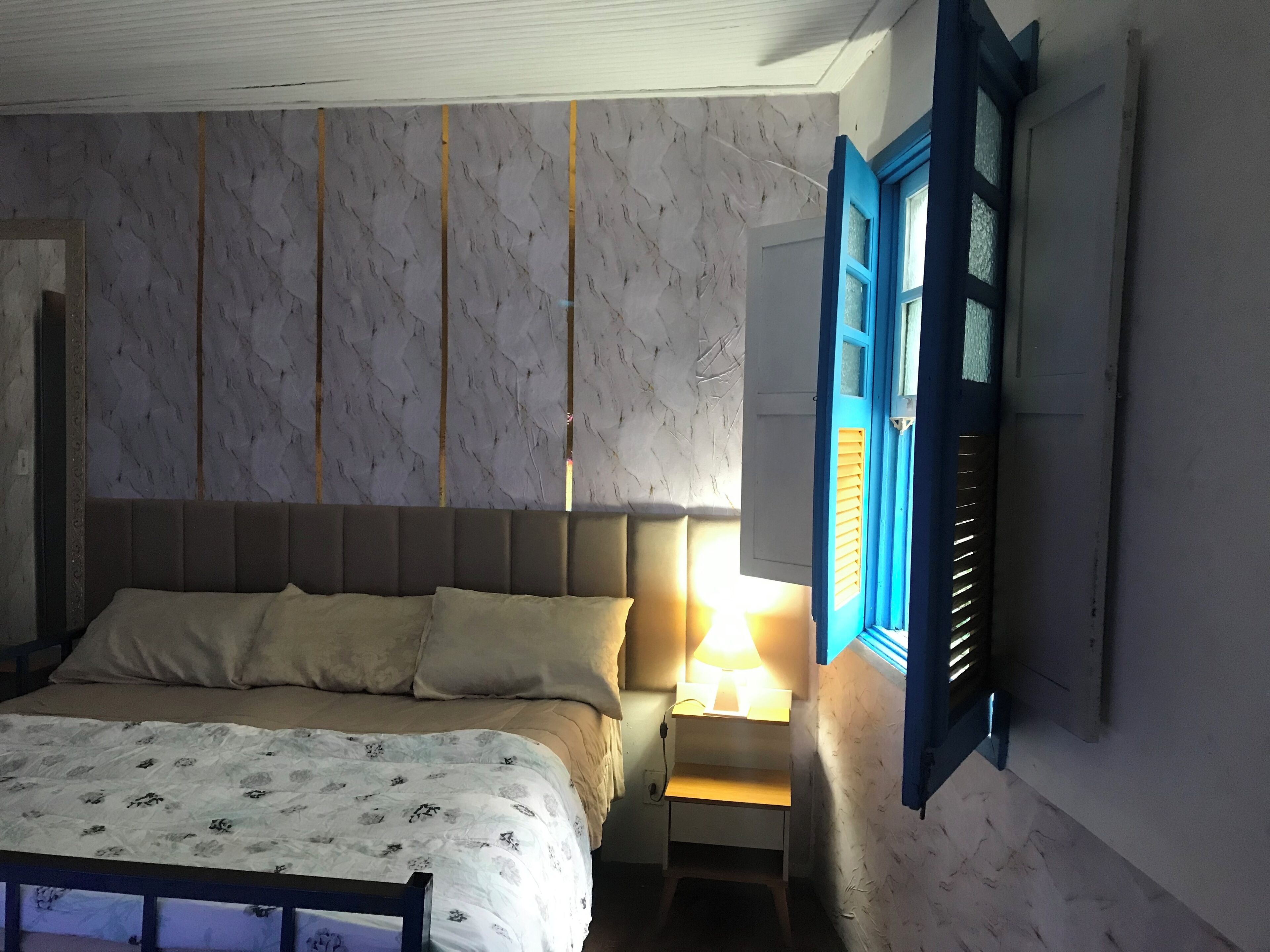 A bedroom with a bed, a bedside table with a lamp, and a window with blue shutters.