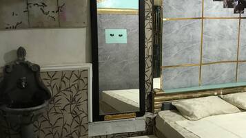 A bedroom with a bed, a large mirror, a wall-mounted radiator, a small sink, and a window with a view of the outside.