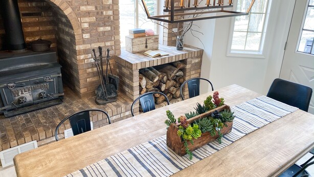 A cozy dining area with a brick fireplace, a wooden table with a striped runner, and a wooden planter with succulents. - The Gathering Place on the Teton River (Rexburg)
