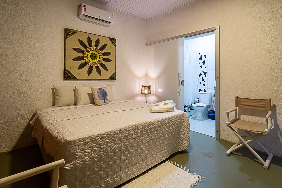 Un dormitorio con cama, una silla, un cuadro y un baño visible a través de una puerta abierta.