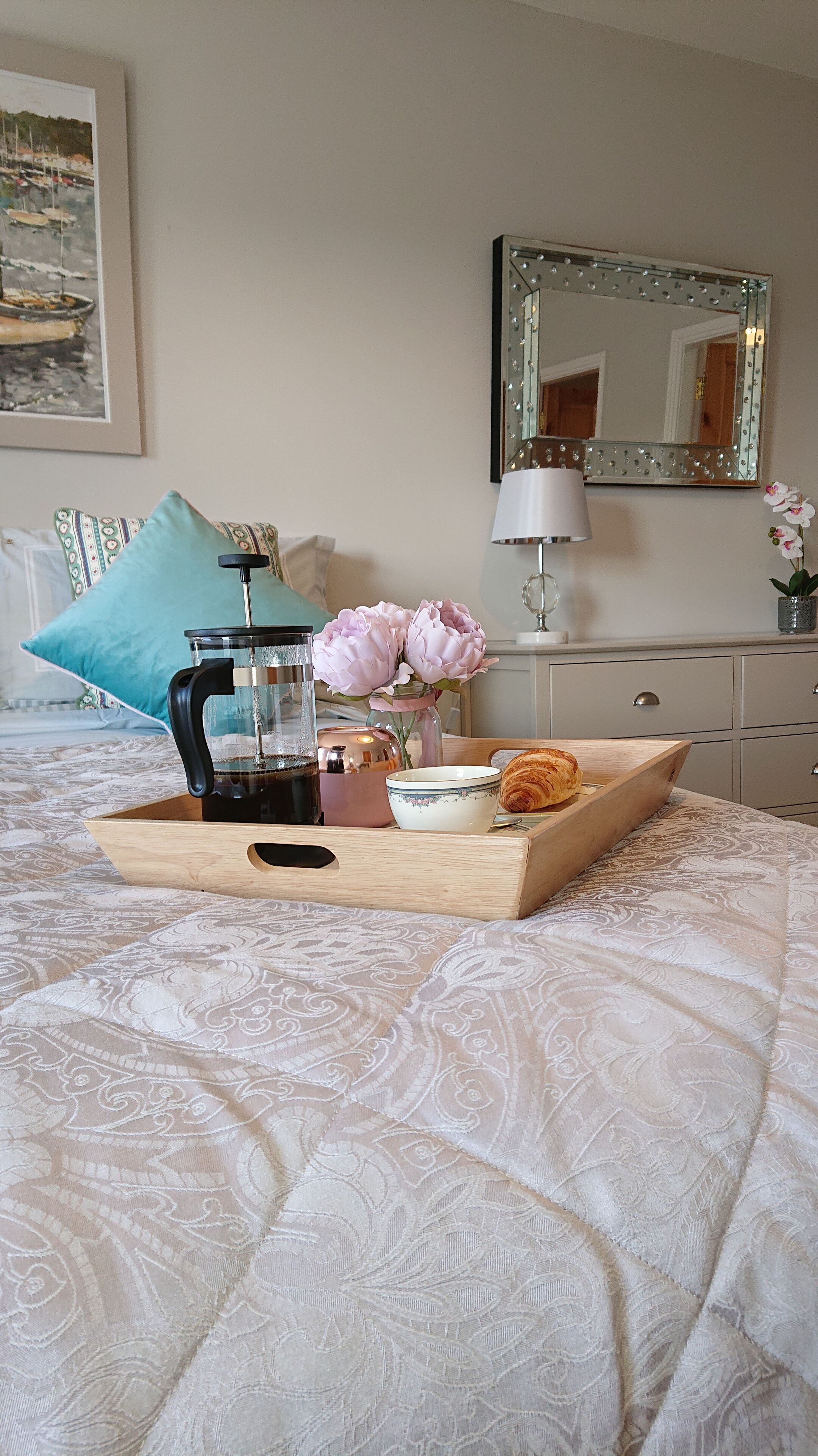 A neatly made breakfast tray on a bed with a patterned bedspread, a coffee maker, a bowl, a croissant, and a vase with flowers.