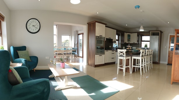 A spacious living area with teal armchairs, a dining table, and a kitchen with white cabinets and stainless steel appliances. - Luxurious and Spacious 5 Bedroom Self-Catering House in Doolin. 3250 Square ft	. (Doolin)
