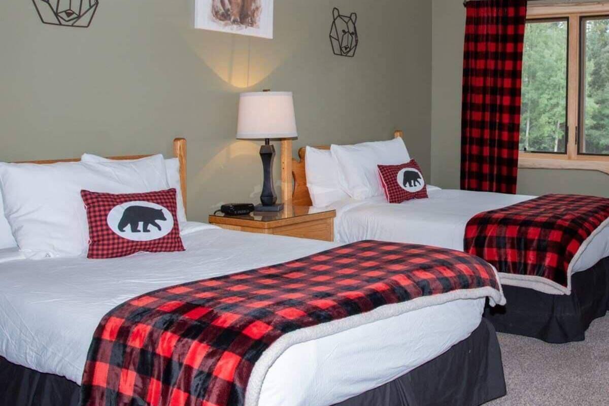 Una habitación con dos camas, cada una con una colcha a cuadros rojo y negro y un cojín con un diseño de oso. Hay una lámpara en una mesita de noche, una ventana con cortina a cuadros rojo y negro, y decoración de pared con temática de osos.