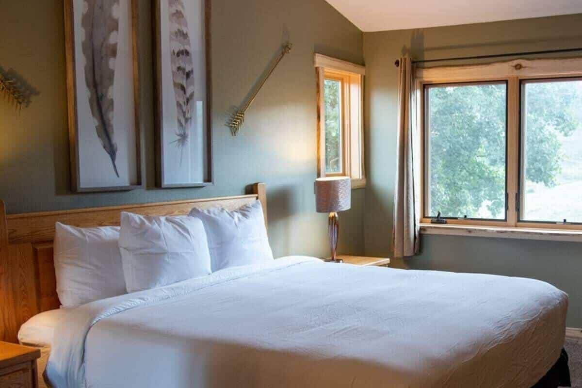 Una cama grande con ropa blanca, un cabecero de madera, una mesita de noche con una lámpara y plumas de pájaro enmarcadas en la pared.