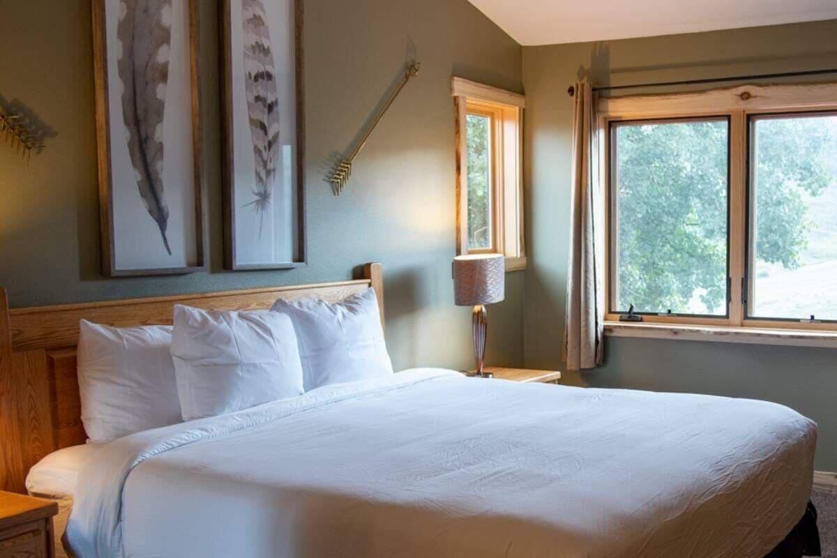 Una cama bien tendida en una habitación con un ventanal grande, una obra de arte enmarcada en la pared y una lámpara de noche.