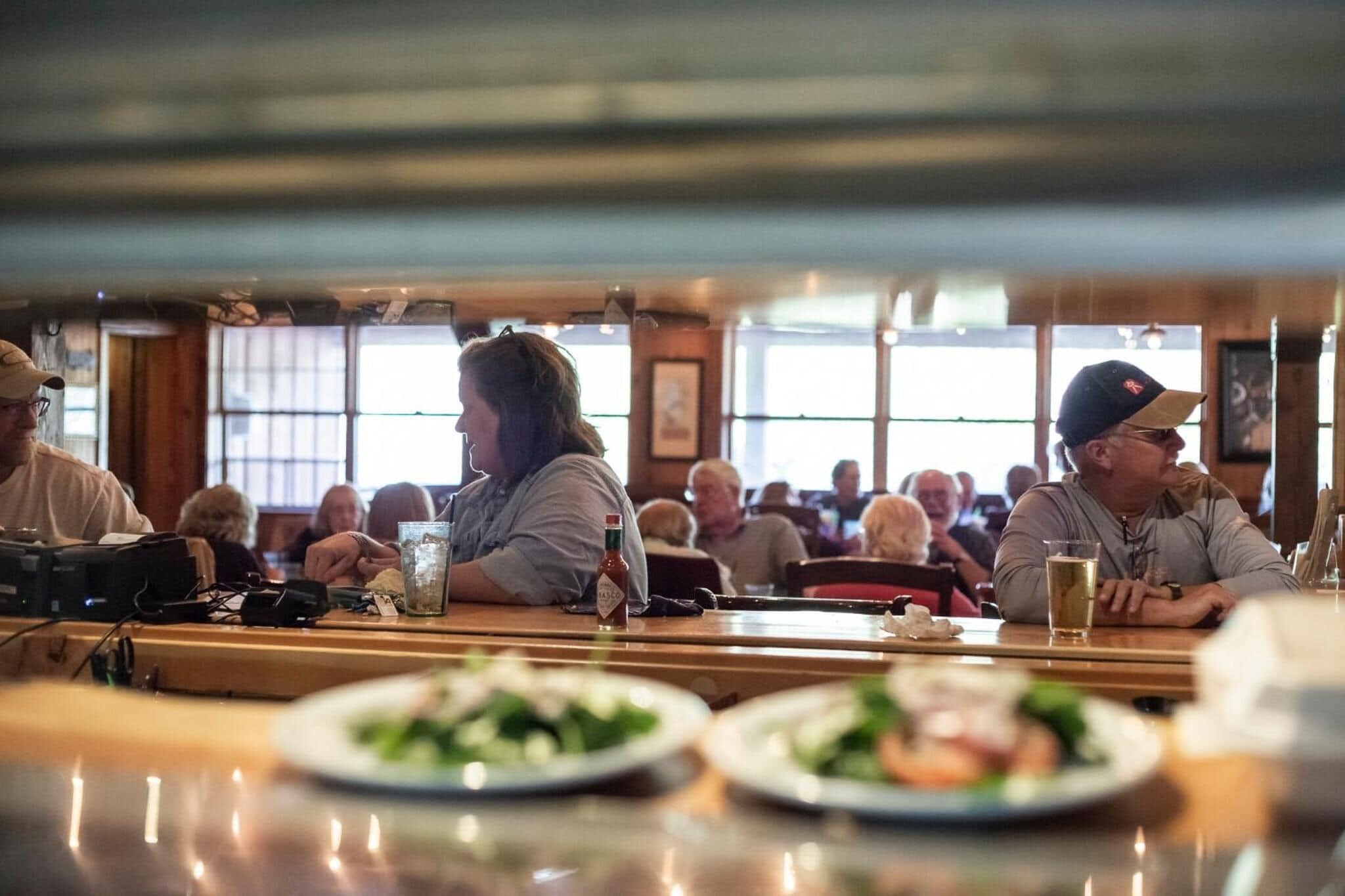 A restaurant interior with wooden tables and chairs, large windows, and patrons dining.