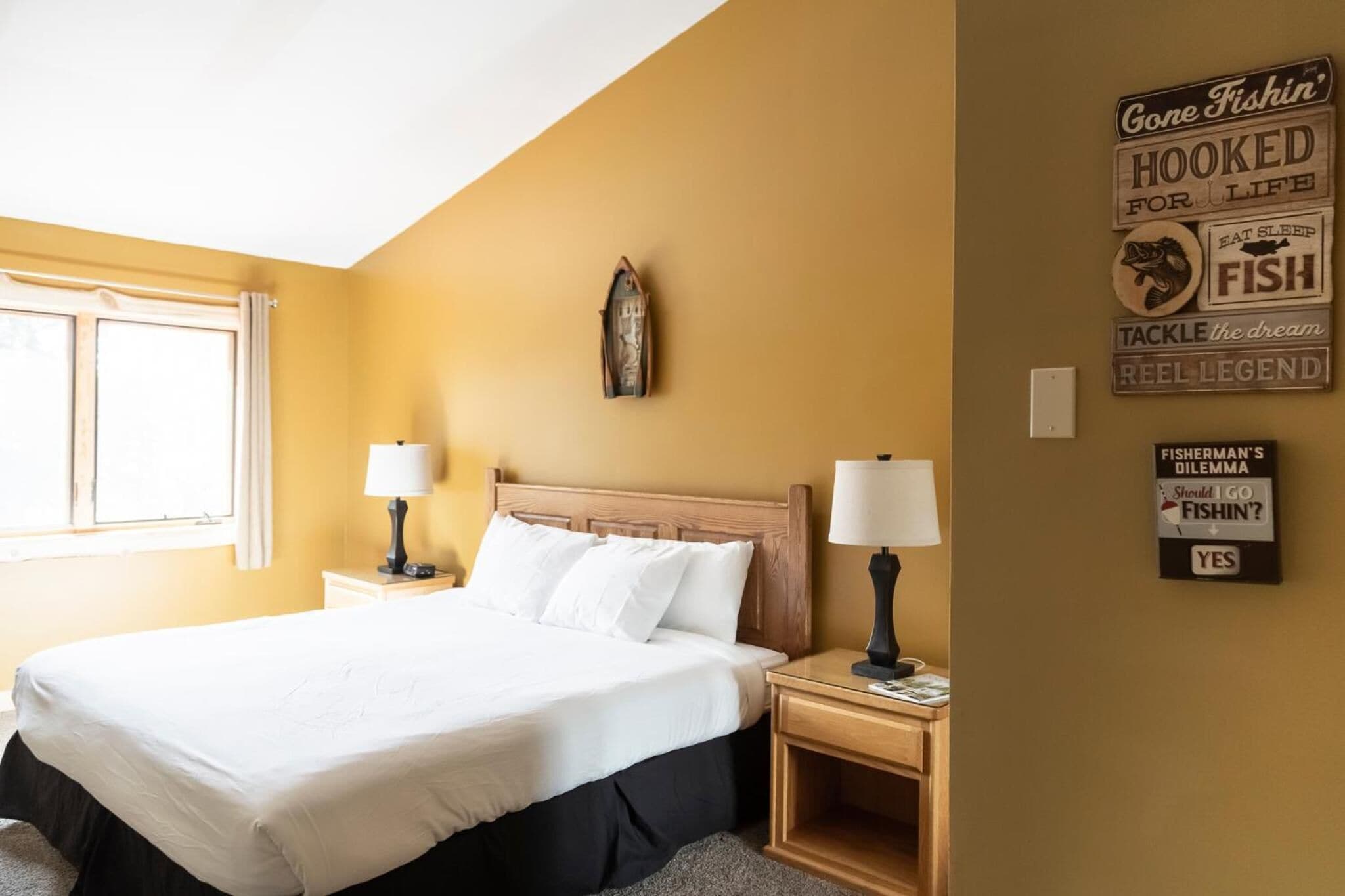 Une chambre d’hôtel comprenant un lit, deux tables de chevet avec des lampes, une fenêtre et des éléments de décoration murale, notamment des panneaux avec des expressions liées à la pêche.