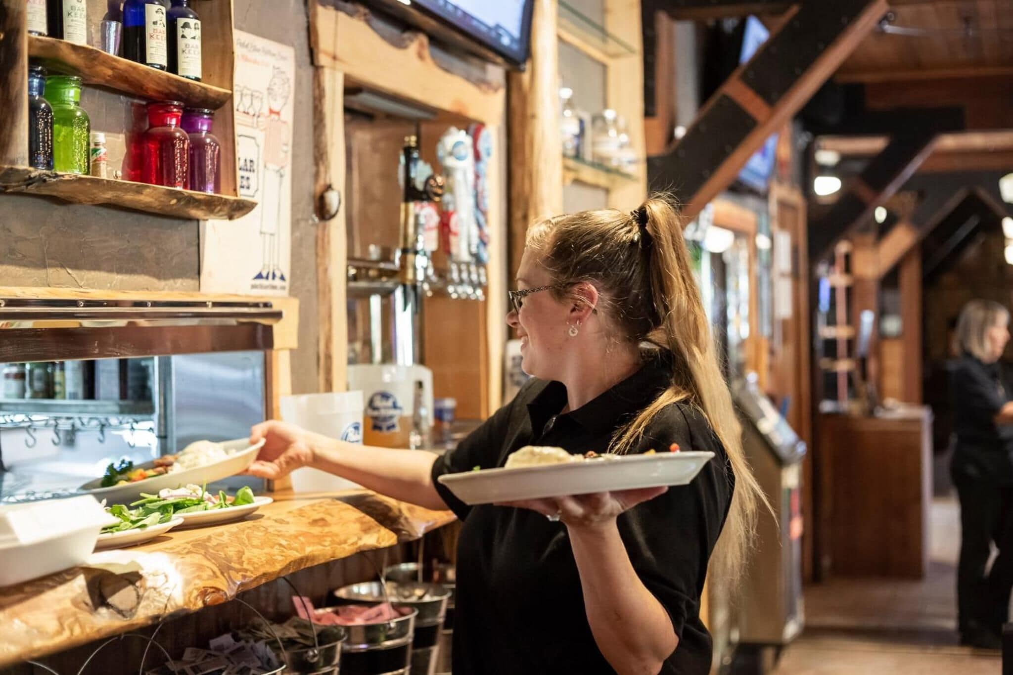 Una persona sirviendo comida en un restaurante con interiores de madera y estantes llenos de botellas.