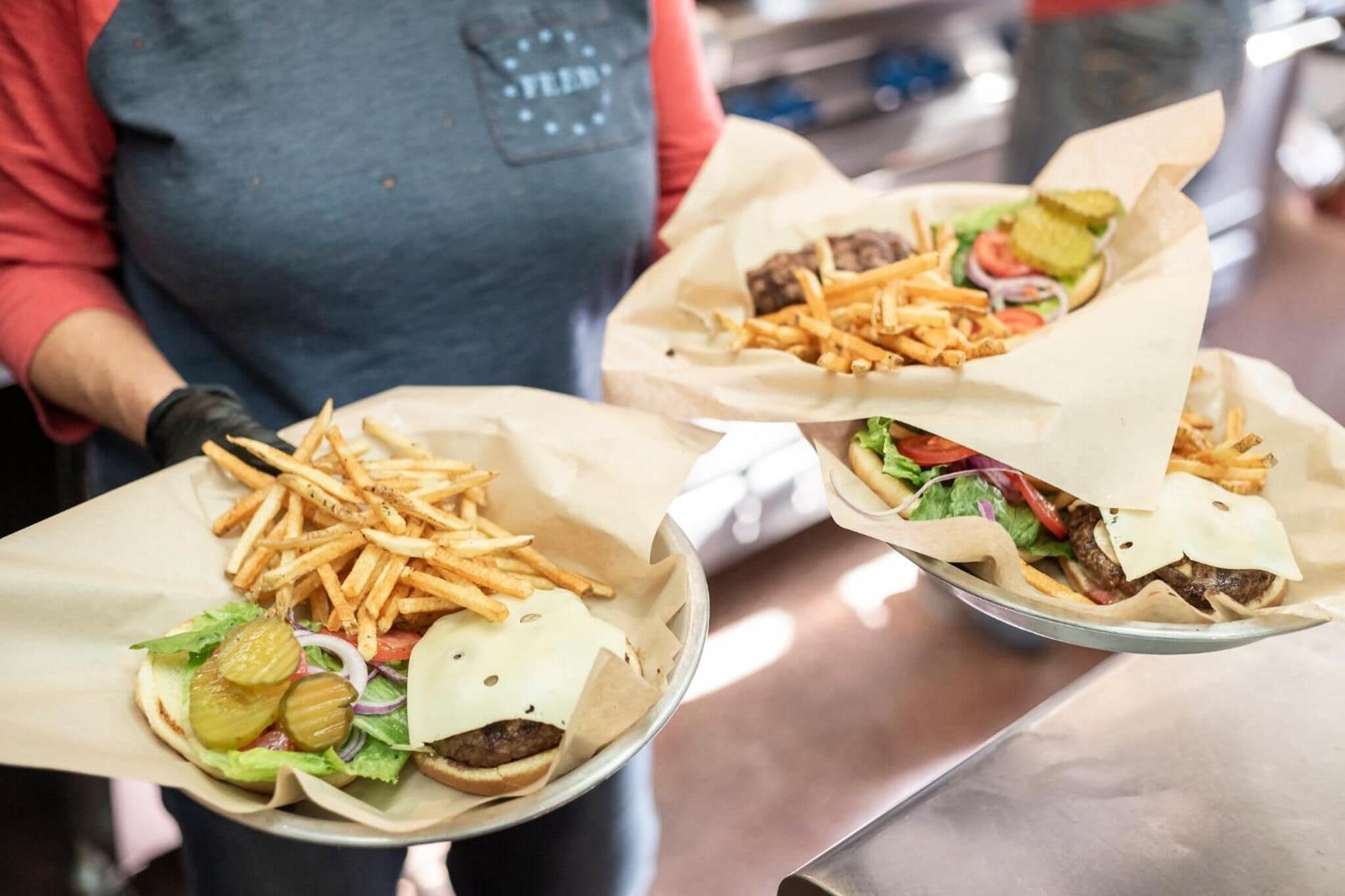 Une personne tenant deux hamburgers emballés, accompagnés de frites et de cornichons.