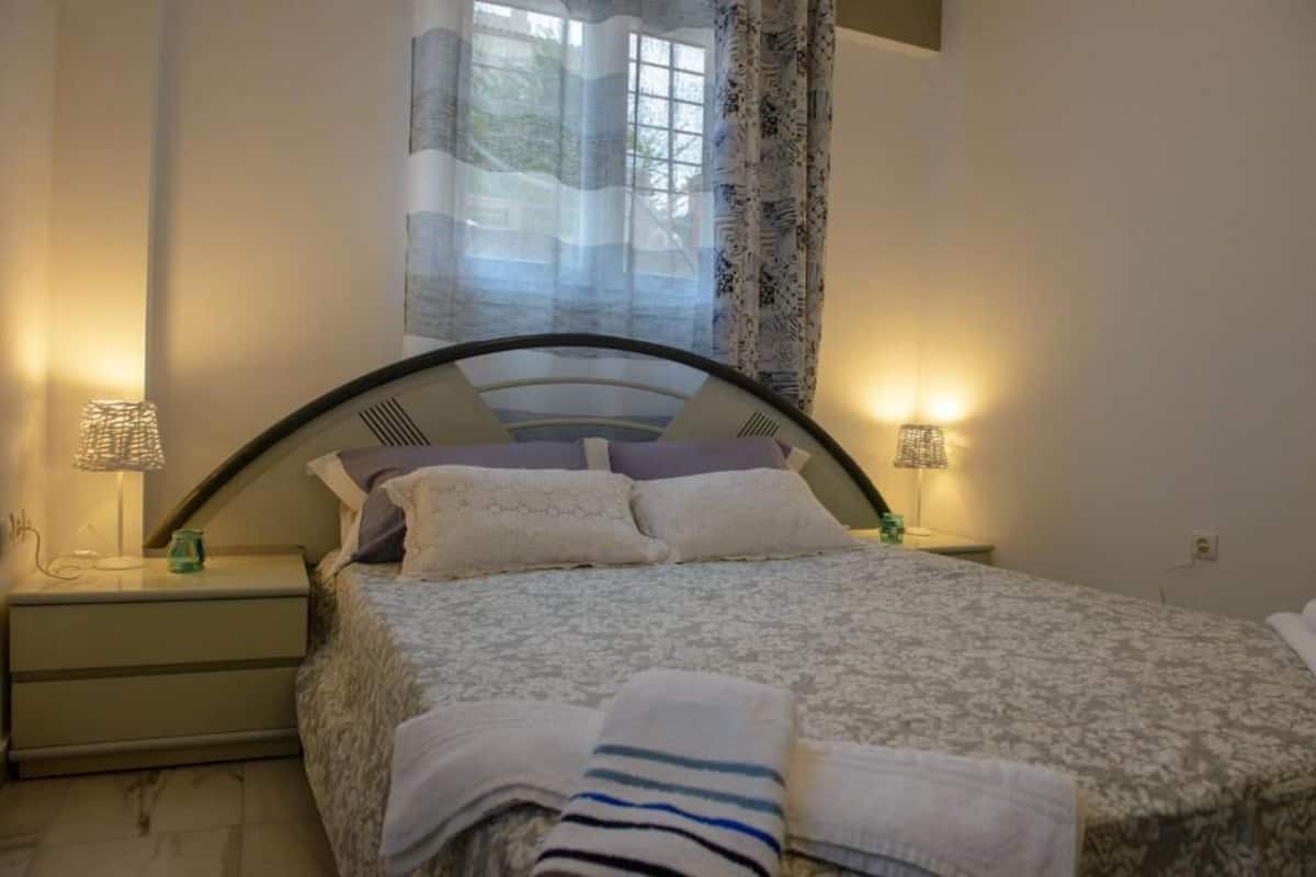 A neatly made bed with a patterned bedspread and pillows, two bedside lamps, and a window with sheer curtains.
