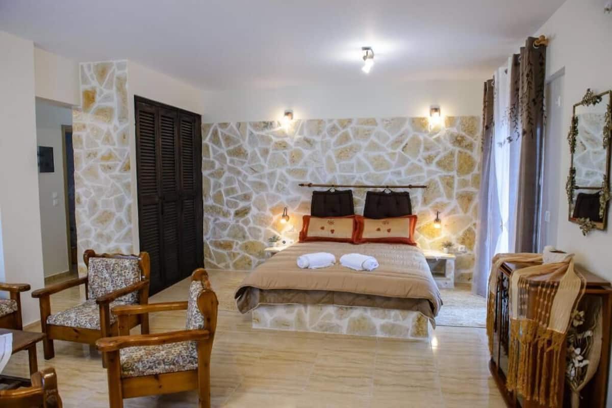 A bedroom with a stone wall, a bed with pillows, a bedside table, and two chairs.