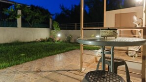 Un patio al aire libre con una mesa redonda de metal y un taburete, rodeado por un muro bajo y un jardín.