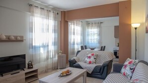 A modern living room with a sofa, a coffee table, a TV, and a kitchen area in the background. - Artemissoula (Spata-Artemida)