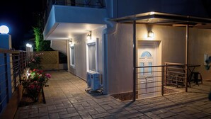 A nighttime view of a building's exterior with a paved entrance, a small garden with flowers, and a security gate. - Artemissoula (Spata-Artemida)