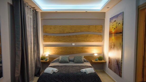 A modern bedroom with a wooden headboard, a large bed with patterned bedding, two bedside tables with lamps, and a large wall painting. - Artemissoula (Spata-Artemida)