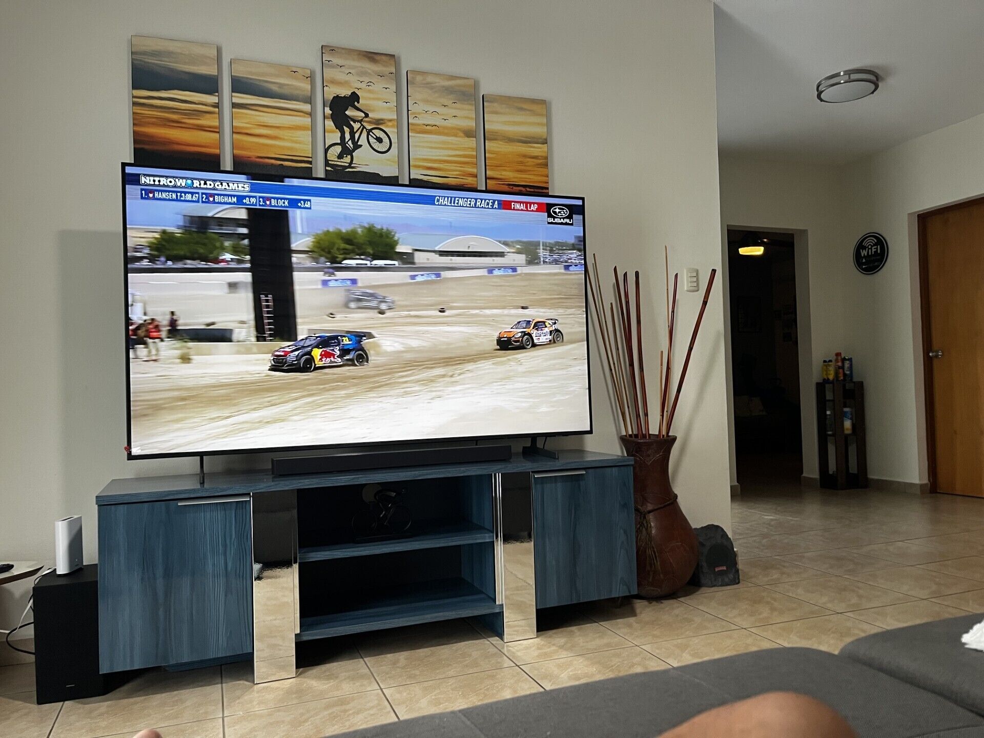 A flat-screen TV mounted on a stand with a racing game displayed, a vase with sticks, and a WiFi sign on the wall.