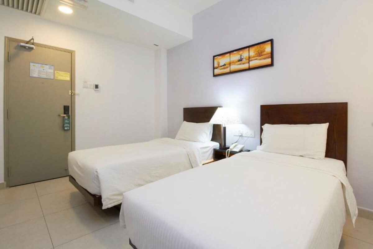 Habitación de hotel con dos camas individuales, una puerta con un letrero, una mesita de noche con una lámpara y un cuadro en la pared.