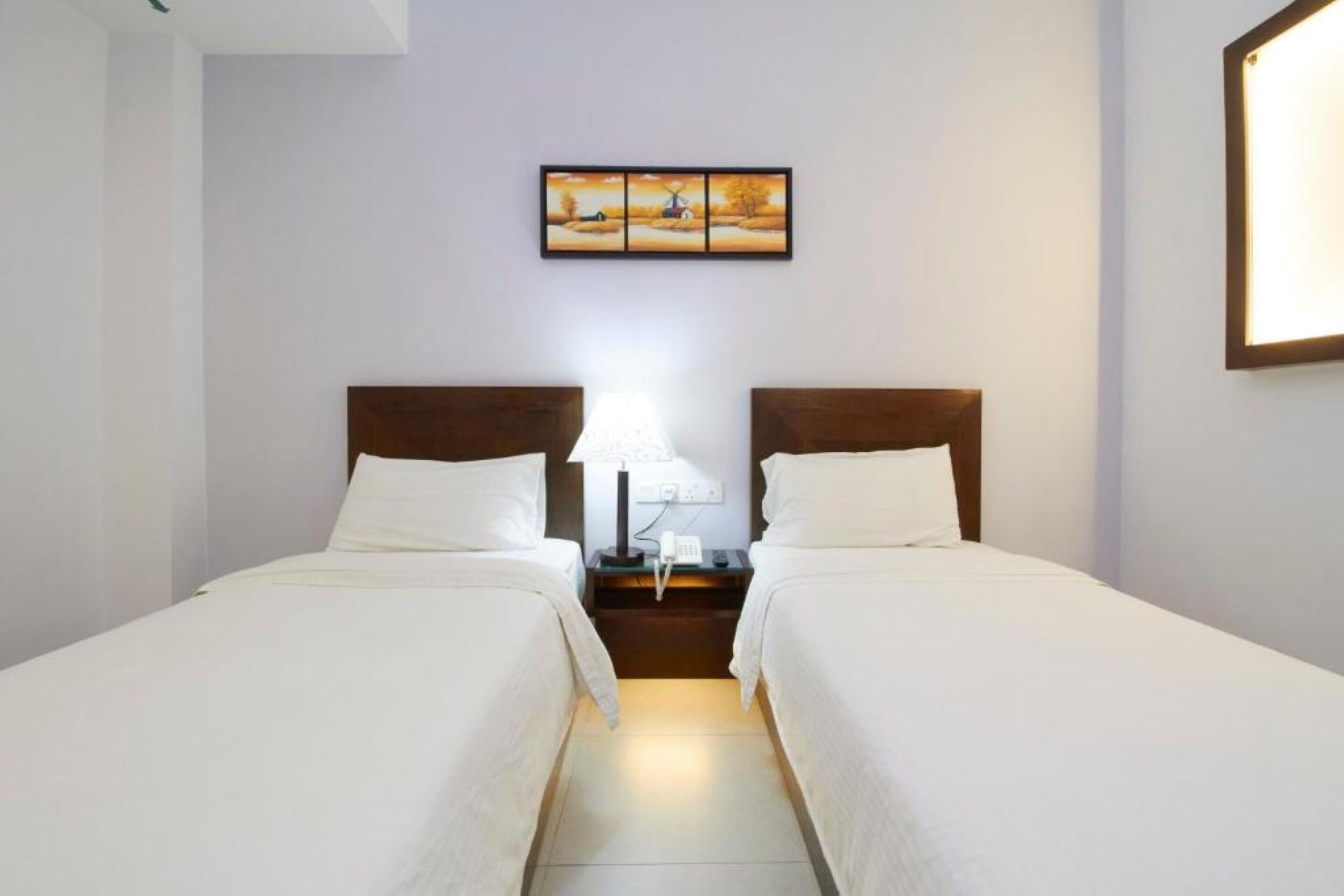 A hotel room with two single beds, a nightstand with a lamp, and a framed picture on the wall.