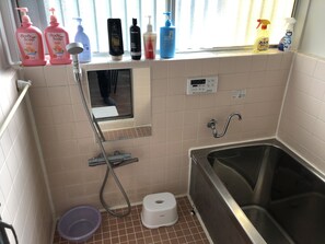 A small bathroom with a shower, toilet, and sink. There are various toiletries on the windowsill, including body soap, shampoo, and a spray bottle.