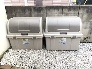 Two gray storage bins with labels, placed on gravel against a brick wall.