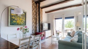 A cozy living room with a dining area, a TV, and a large window with a view of palm trees. - Casa Tossa – Pool, Sea Views & A/C (6 pax) (Tossa De Mar)