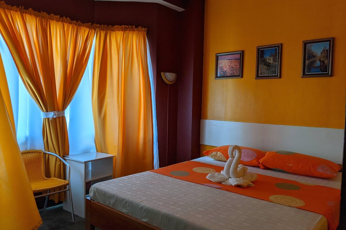 Ein Hotelzimmer mit einem Bett, orangefarbenen Vorhängen, einem kleinen Tisch und zwei gerahmten Bildern an der Wand.