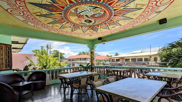 Uma área externa com mesas e cadeiras, um mural colorido no teto e vista para prédios e árvores ao fundo.
