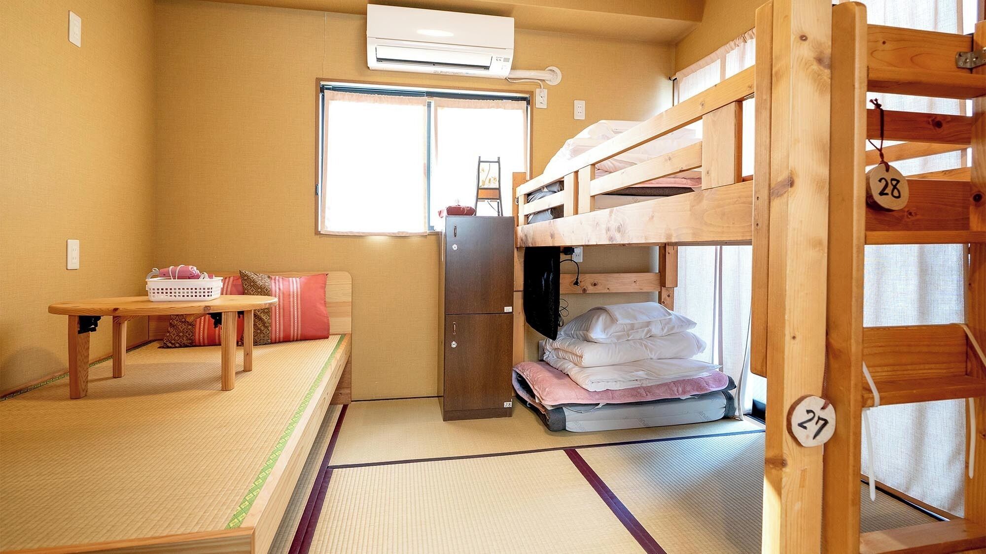 A dormitory room with bunk beds, a small table, a refrigerator, and a window.