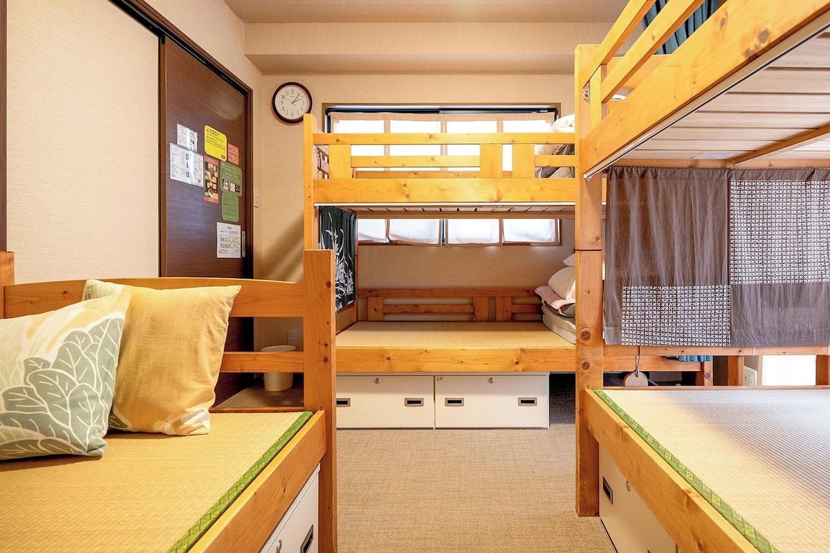 Une chambre avec un lit superposé, un plancher en bois, une horloge au mur et une fenêtre avec un rideau.