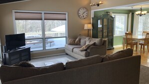 A living room with a large window, a television on a cabinet, a sofa, and a dining area with chairs and a table.